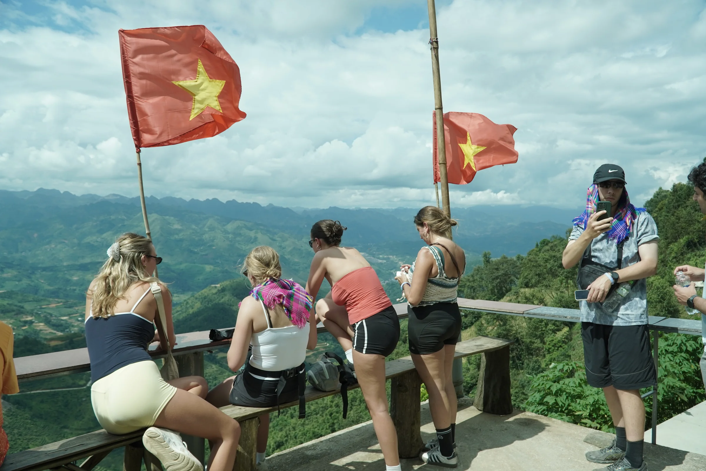 Travellers at a railed viewpoint above a wide green Ha Giang valley, two Vietnamese flags flying overhead