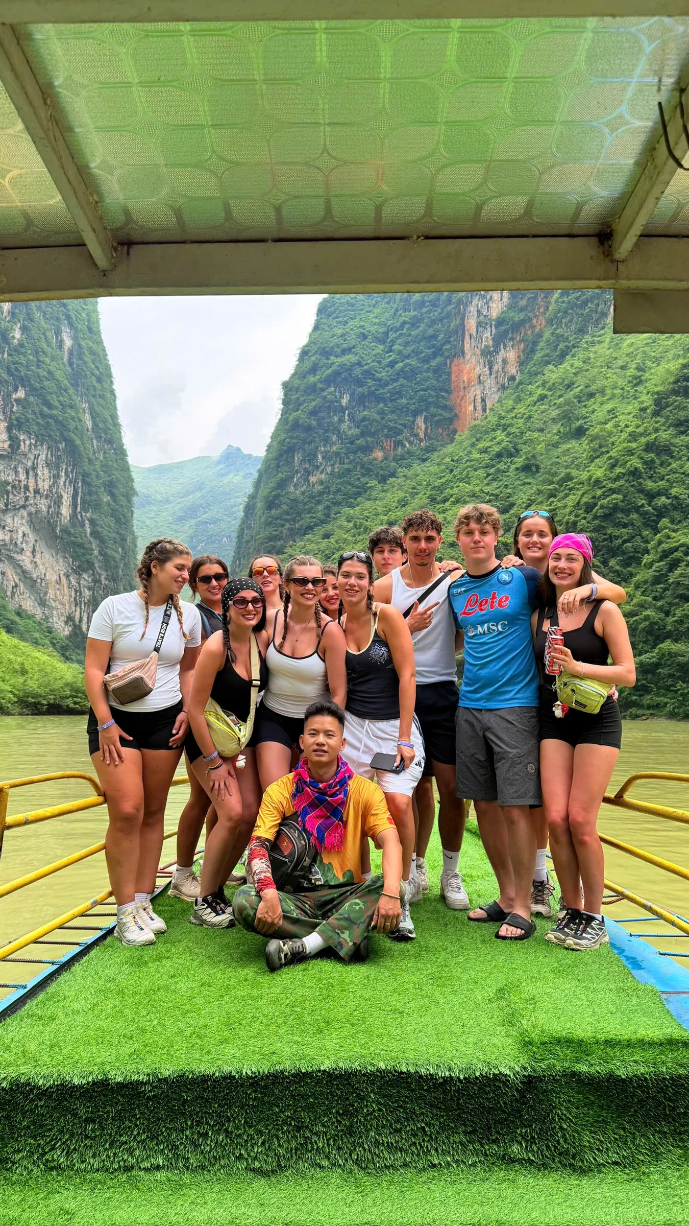 Travellers on the green deck of a boat between near-vertical limestone cliffs on the Nho Que river