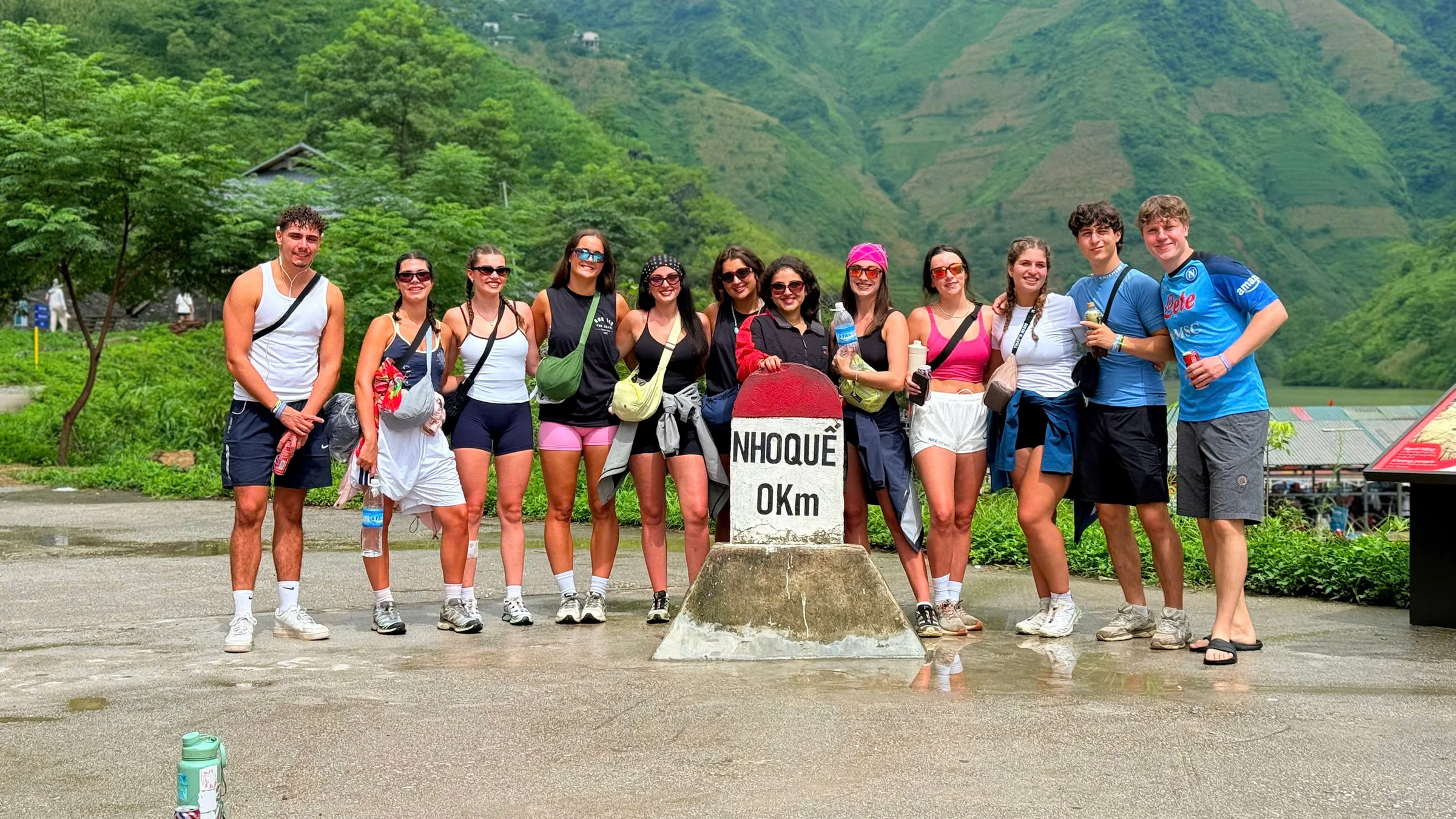 A group of travellers standing around a red and white stone kilometre marker reading NHO QUE 0Km, with green mountainsides and the river behind