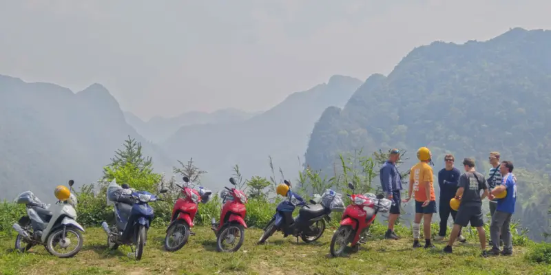 A line of rental motorbikes parked on a grass verge with travellers beside them, hazy karst mountains behind, Ha Giang, April 2026