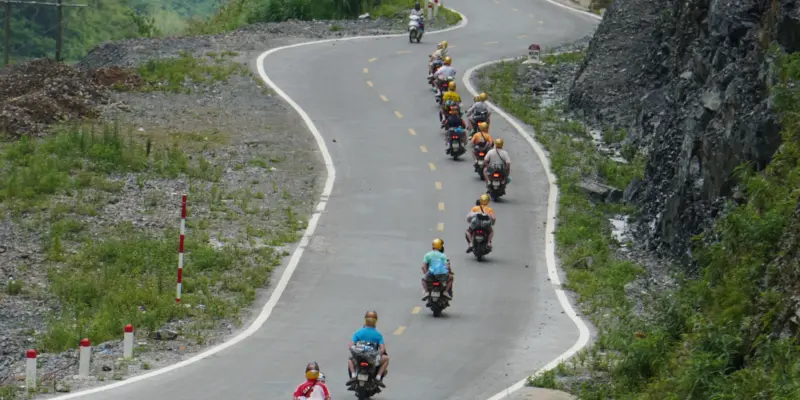 A long line of motorbike riders strung out along an S-curve of paved mountain road in Ha Giang, seen from above