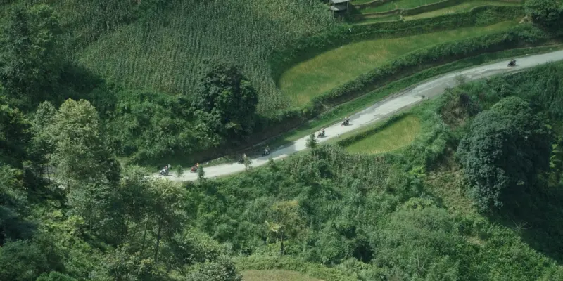 A line of motorbikes riding spaced apart along a narrow paved road cut into a green hillside above terraced fields, Ha Giang, July 2026