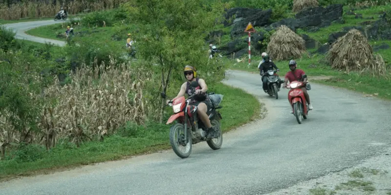 A rider on a red trail bike coming down a single-lane road on the Ha Giang Loop, with more riders following behind through green karst hills