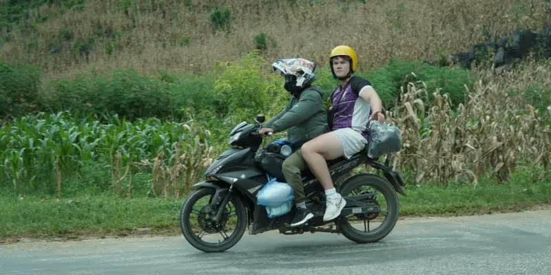 Two travellers riding two-up on a semi-automatic motorbike along a quiet paved road through green hills in Ha Giang