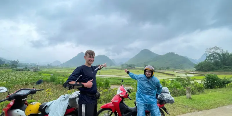 A rider in a blue rain poncho standing beside a red rental scooter on a grass verge above flooded rice terraces in Ha Giang, June 2026