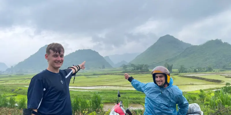 Two riders beside a motorbike in wet-season conditions on the Ha Giang Loop, one in a blue poncho, luggage wrapped in plastic, flooded rice terraces and low cloud behind