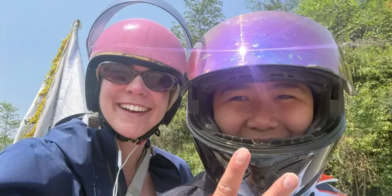 A Ha Giang Loop easy rider and his passenger in helmets at a roadside stop, the rider giving a peace sign to the camera