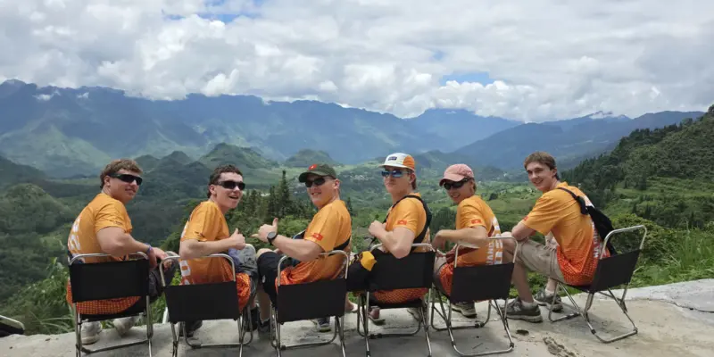 Travellers seated in a row at a roadside viewpoint in Ha Giang with mountains behind, July 2026
