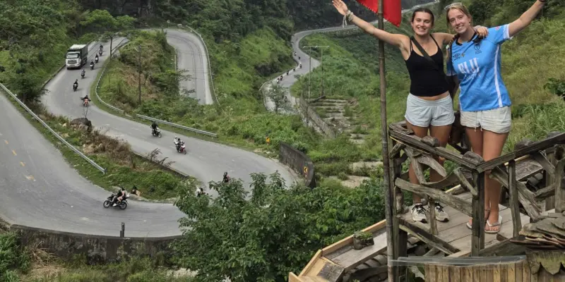Switchbacks of a mountain road seen from above, with motorbikes spread along it and an articulated truck coming down one of the hairpins