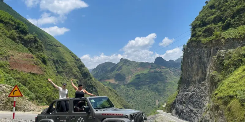 A narrow paved road curving between steep karst walls in Ha Giang with a yellow sharp-bend warning sign at the roadside, June 2026