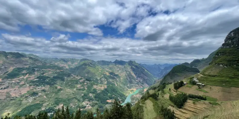 A deep valley in Ha Giang with terraced fields on the slopes and the Nho Que river far below, heavy cloud overhead, April 2026
