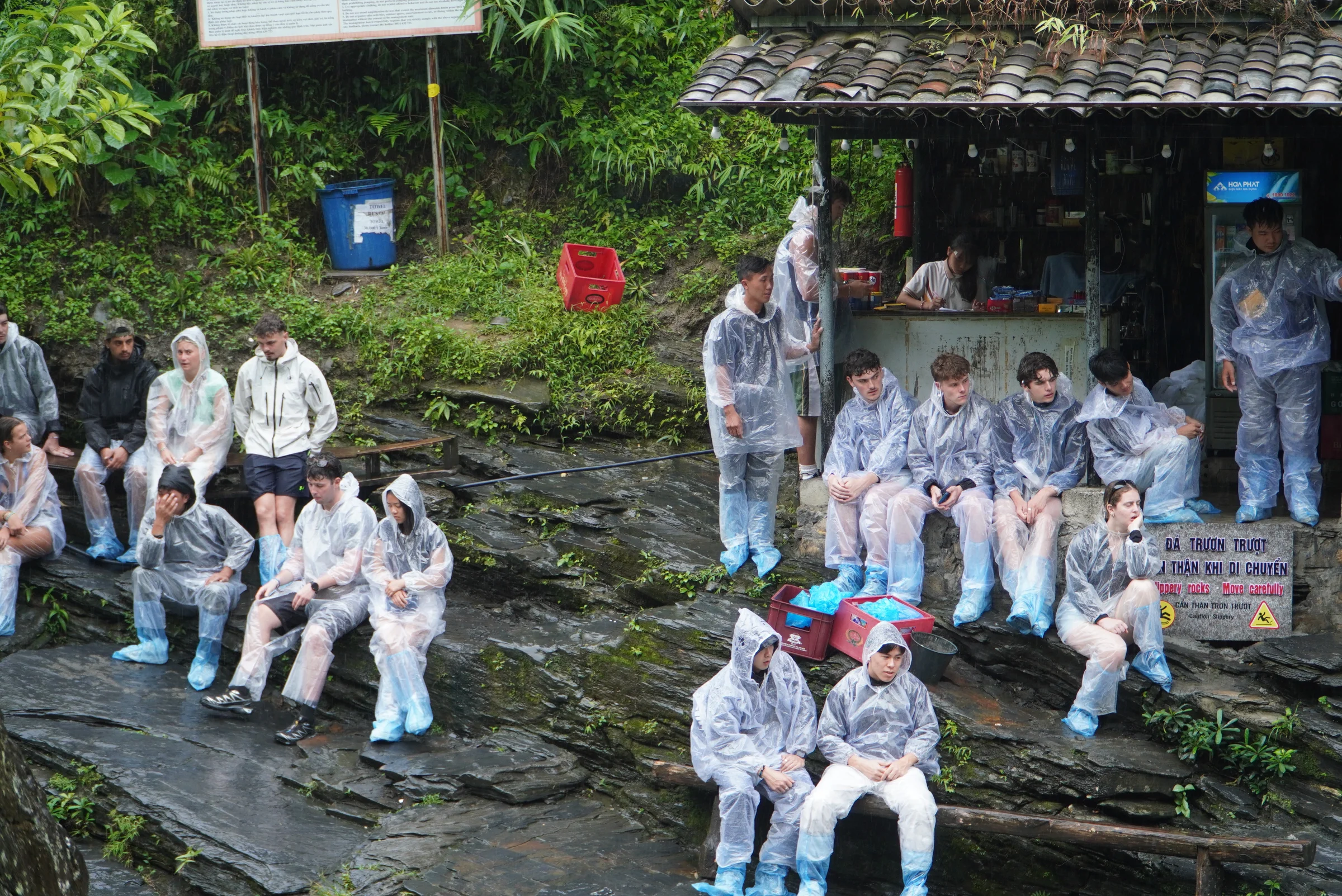 A large group in disposable plastic rain ponchos and blue shoe covers sitting on wet rock beside a small kiosk, next to a bilingual sign warning that the rocks are slippery