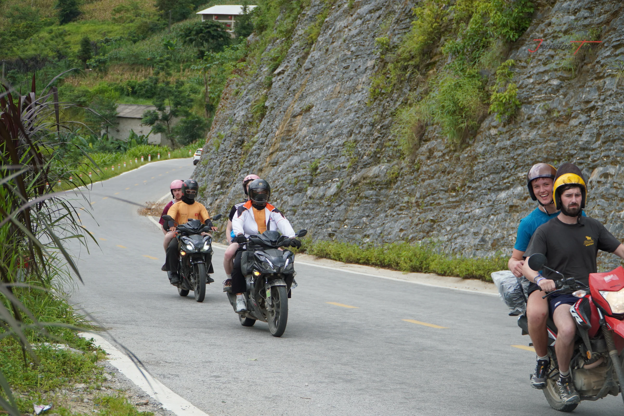 Two riders on motorbikes taking a bend on a mountain road with a cut rock face on one side