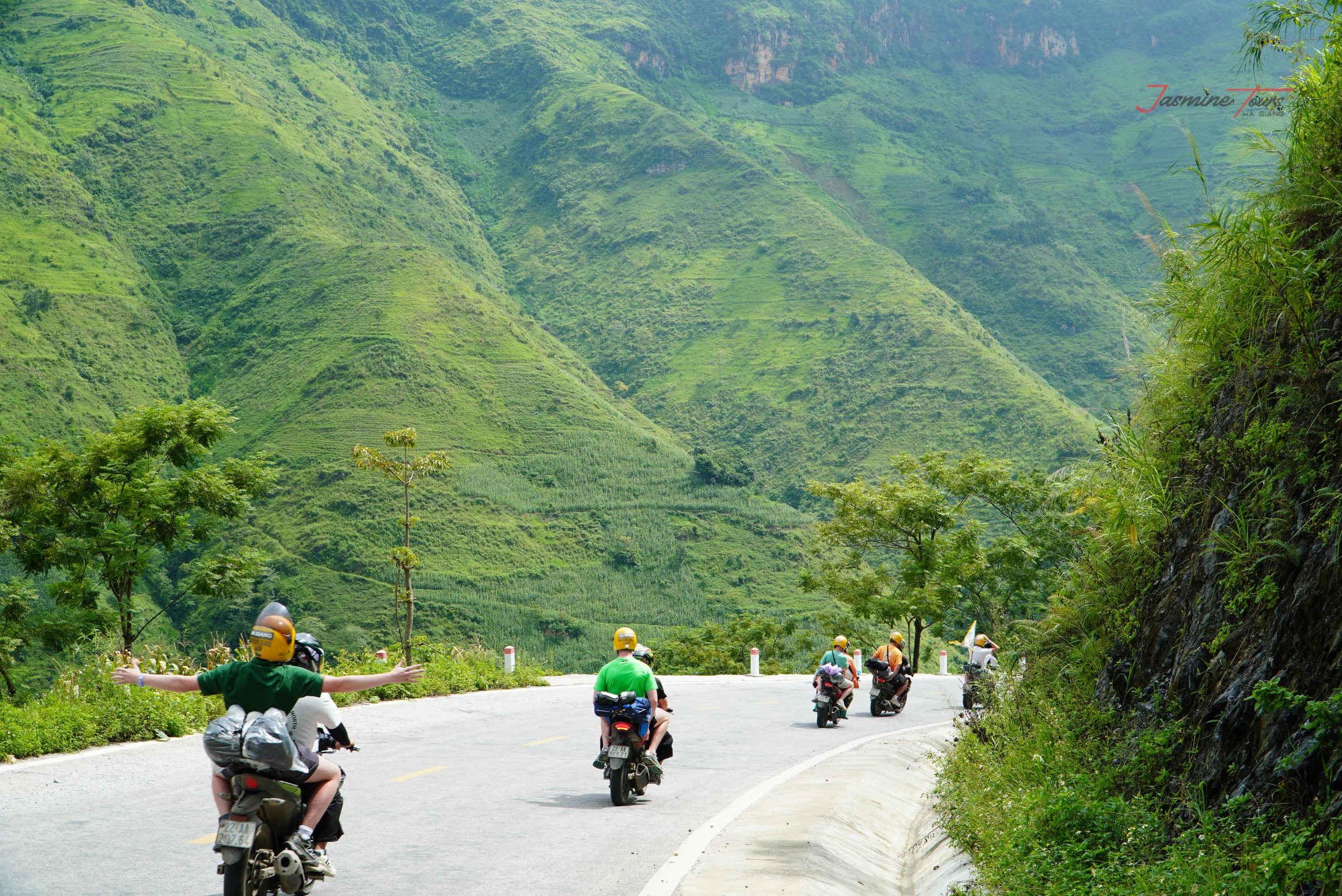 Riders spread out along a smooth paved mountain highway with a kerb, drainage channel and marker posts, green ridges behind