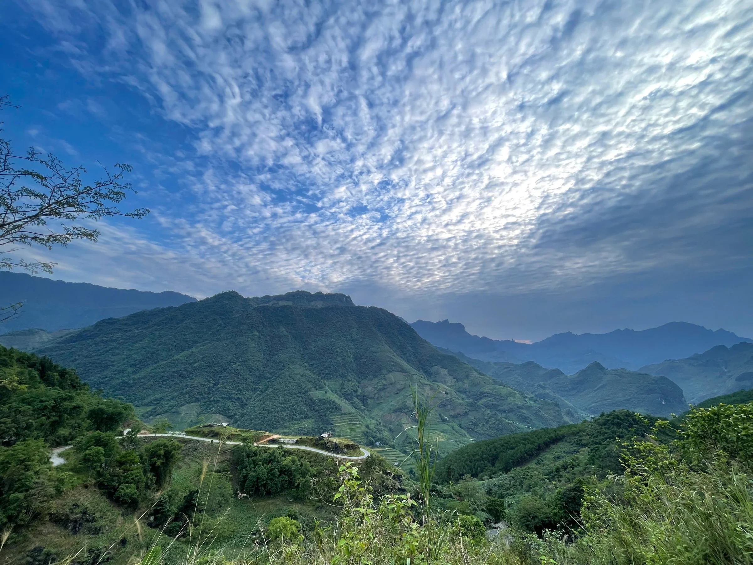 A band of high cloud spread across the sky above layered green mountains and a switchback road in Ha Giang, May 2026
