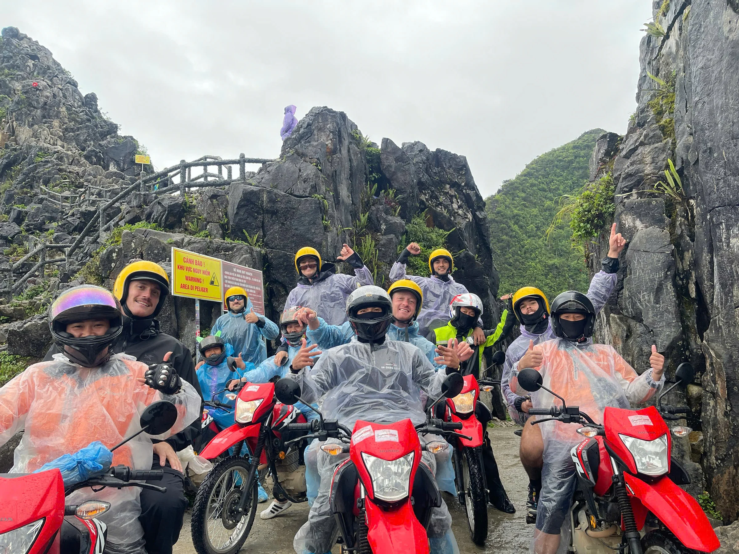 A group of riders in full helmets and rain ponchos beside red trail bikes, next to a yellow Vietnamese and English sign reading area of danger, Ha Giang, May 2026