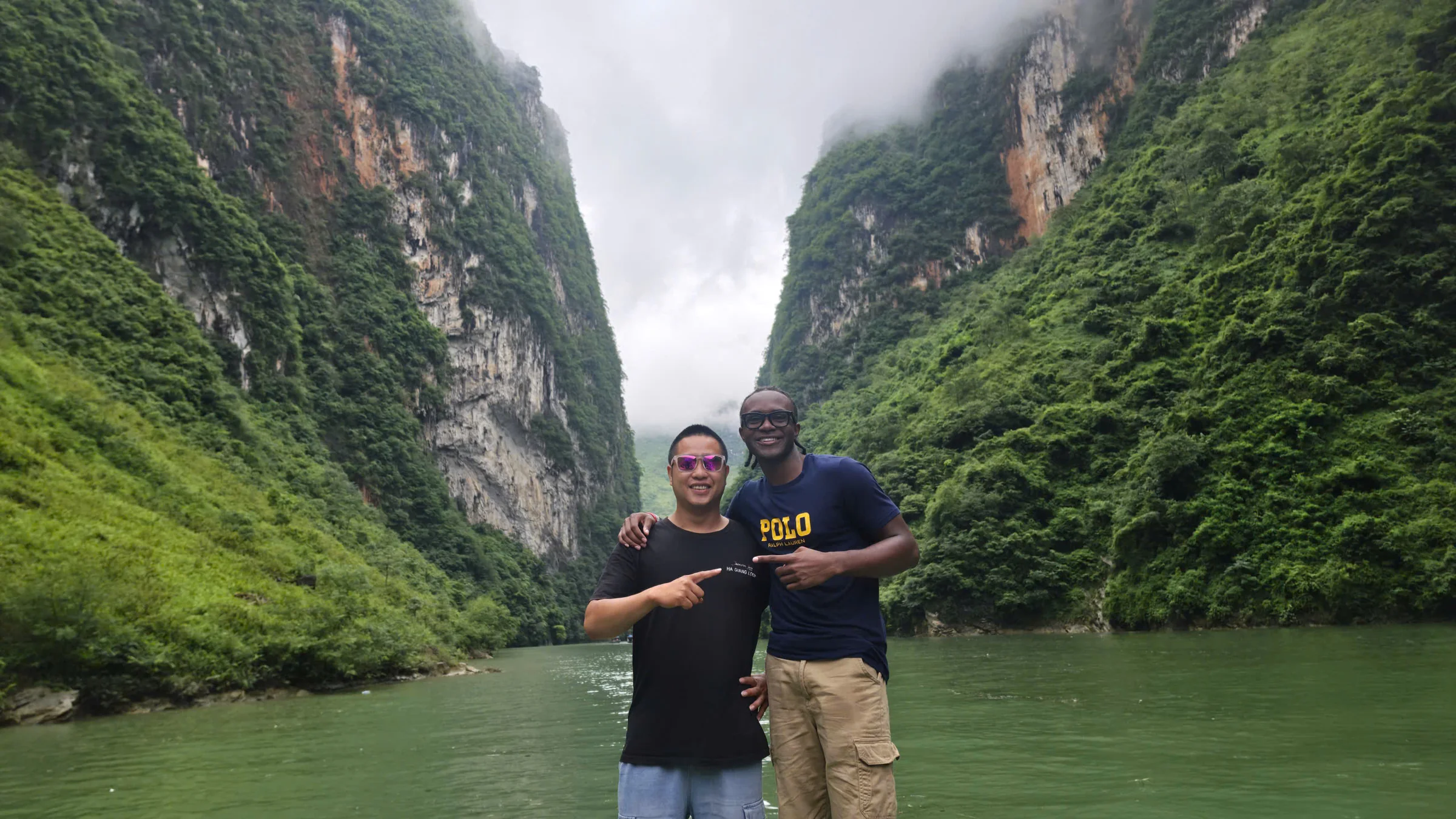 Two travellers at the bottom of the Nho Que gorge with sheer limestone walls rising on both sides and low cloud filling the top of the canyon, July 2026