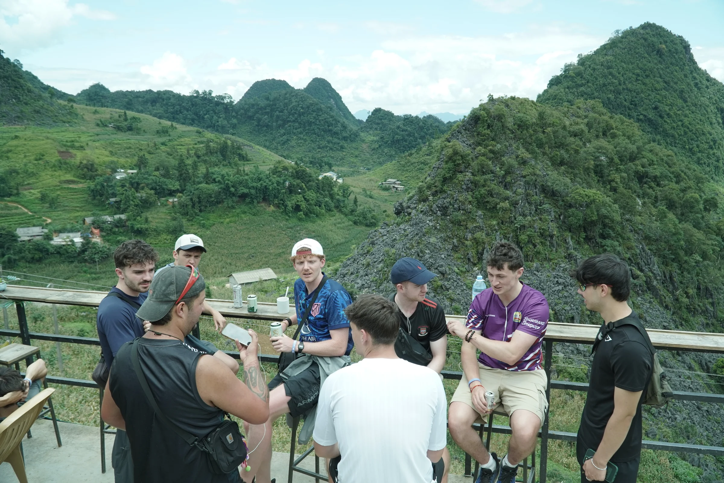A group of travellers standing and sitting at a railed viewpoint above green karst hills in Ha Giang