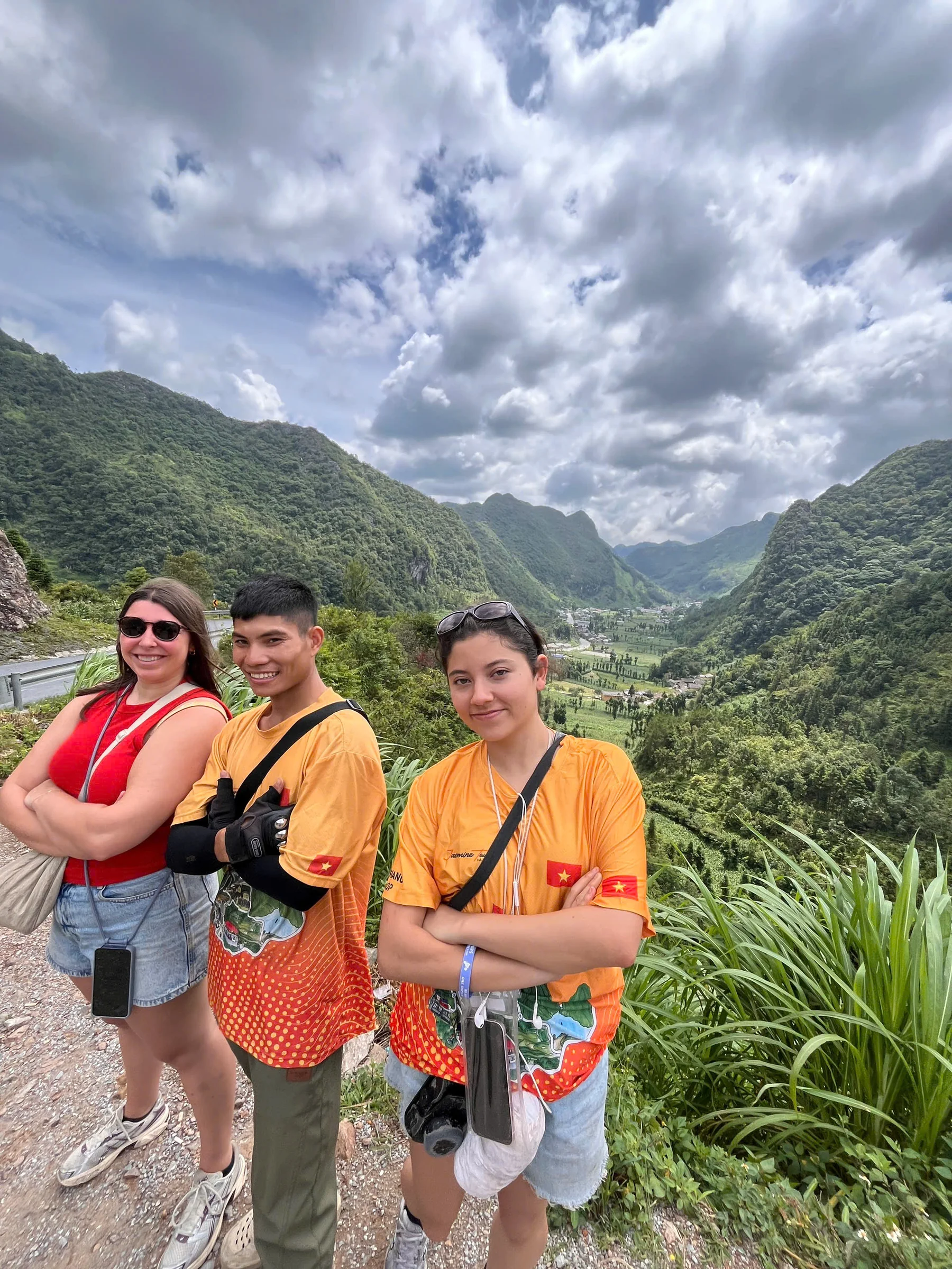 Two local guides in orange tour shirts standing either side of a guest at a Ha Giang viewpoint, a green valley behind