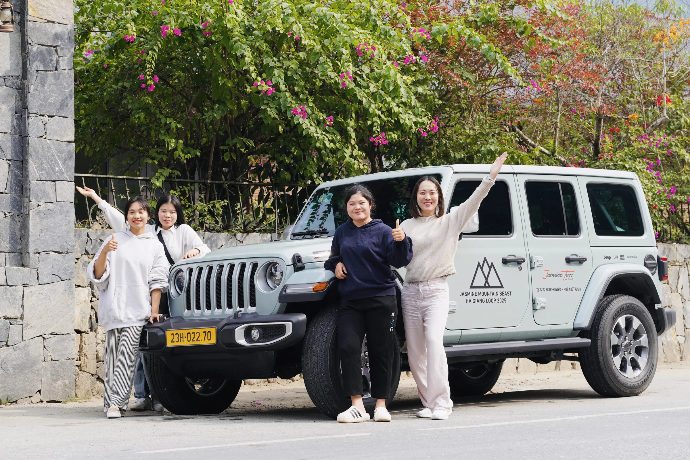 Four travellers standing beside a white Jeep marked Jasmine Mountain Beast Ha Giang Loop 2025, parked under flowering trees on the Ha Giang Loop