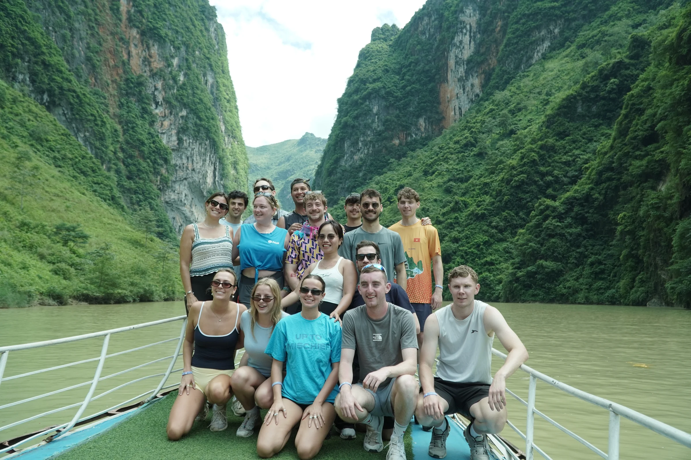 A group of travellers posing on the deck of a boat on the green Nho Que river between towering limestone cliffs in Ha Giang