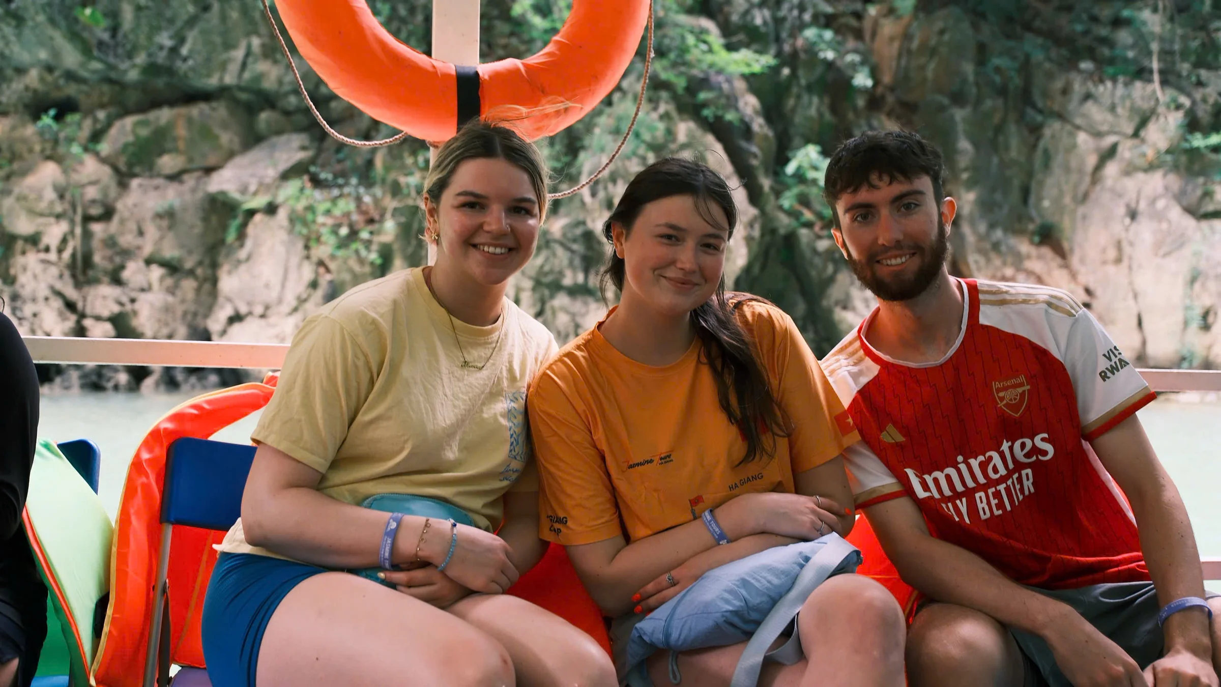 Three travellers seated together on a boat with life jackets and karst cliffs behind, Ha Giang, May 2026