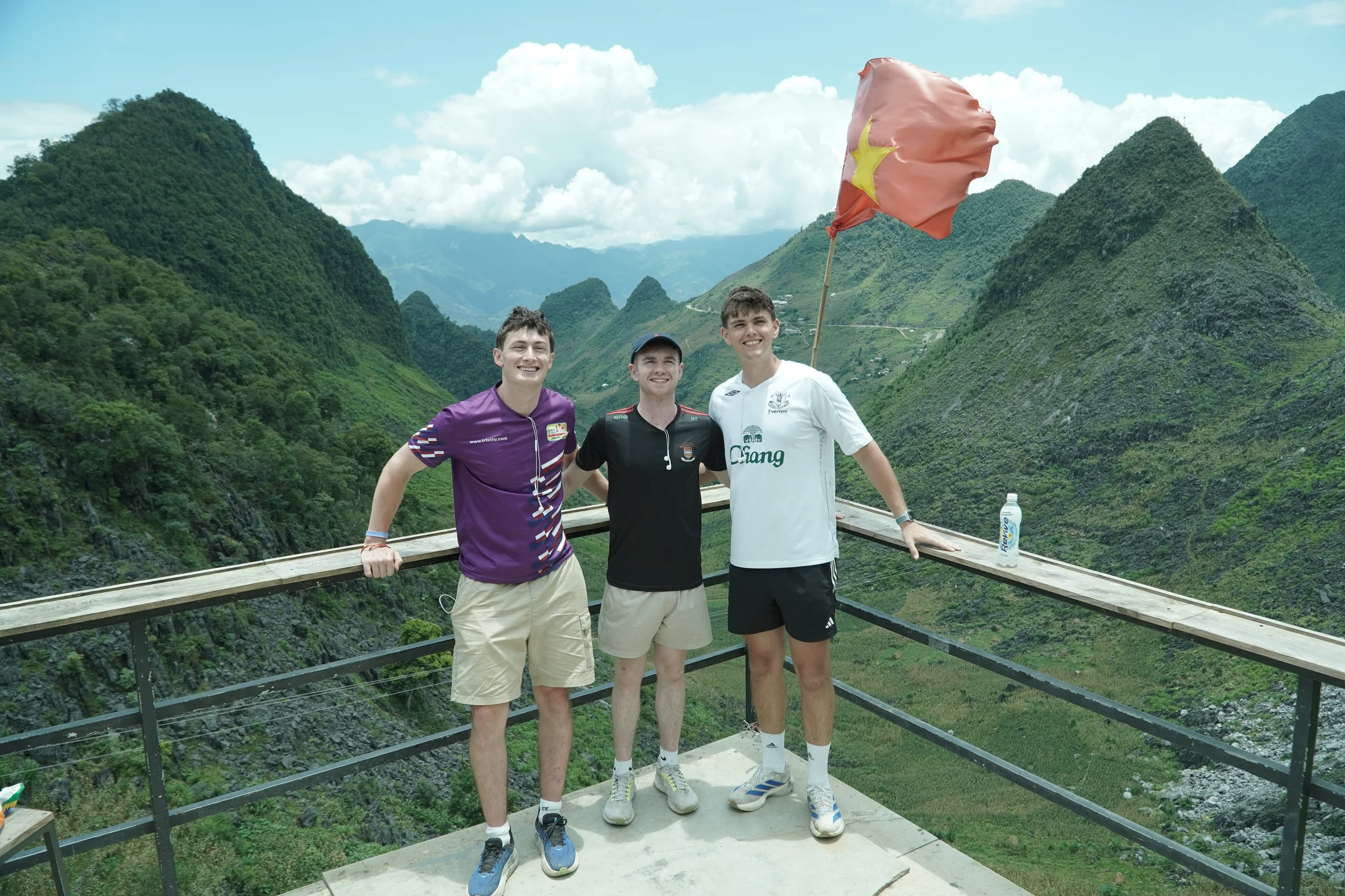 Travellers standing and talking at a mountain viewpoint in Ha Giang, valley and flags behind them