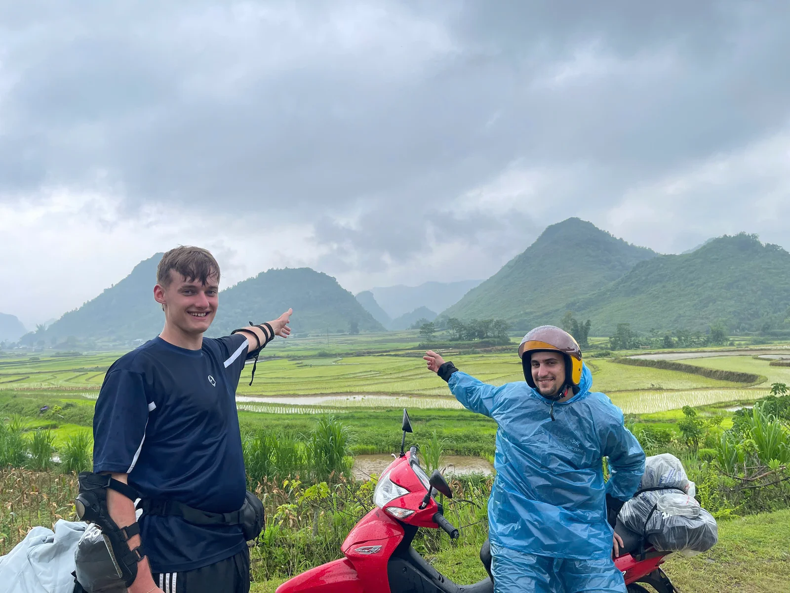 Two travellers standing beside a red rental motorbike on a wet day beside rice terraces on the Ha Giang Loop