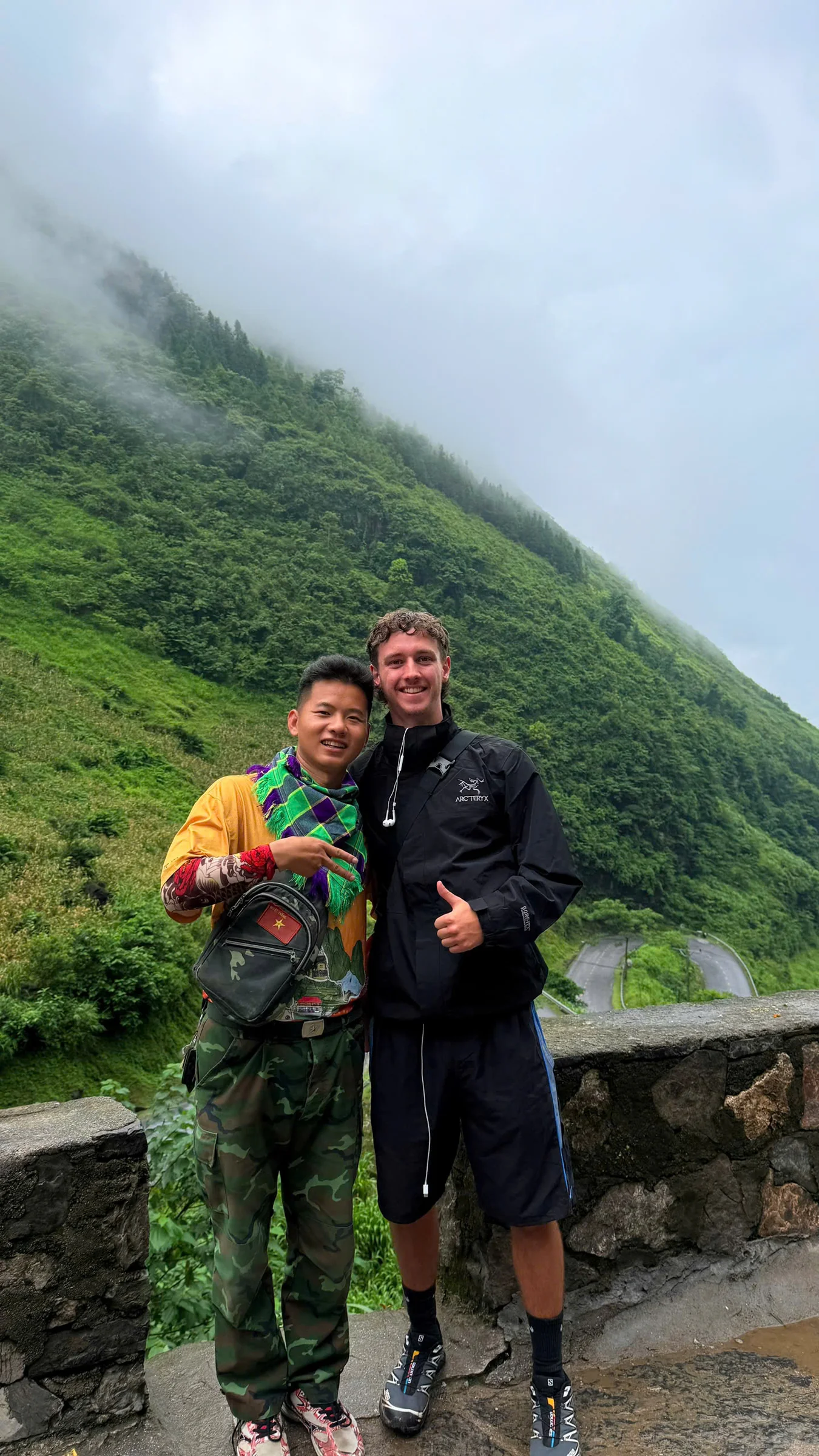 A Ha Giang Loop easy rider with his arm around his passenger at a roadside viewpoint, cloud sitting low on the green ridge behind them