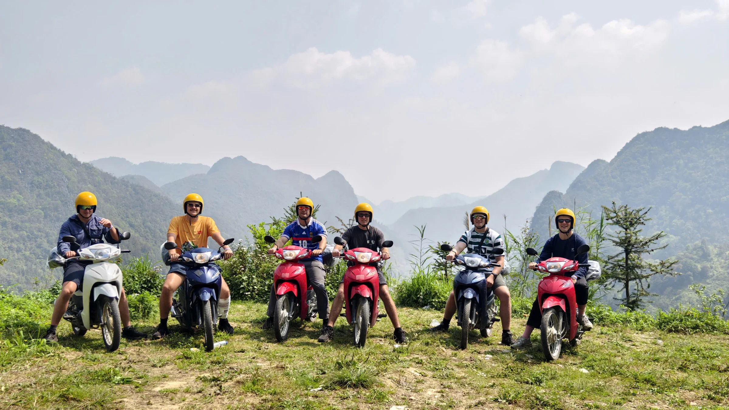 Six travellers sitting on semi-automatic motorbikes in helmets at a viewpoint, layered hazy mountains behind, Ha Giang, April 2026