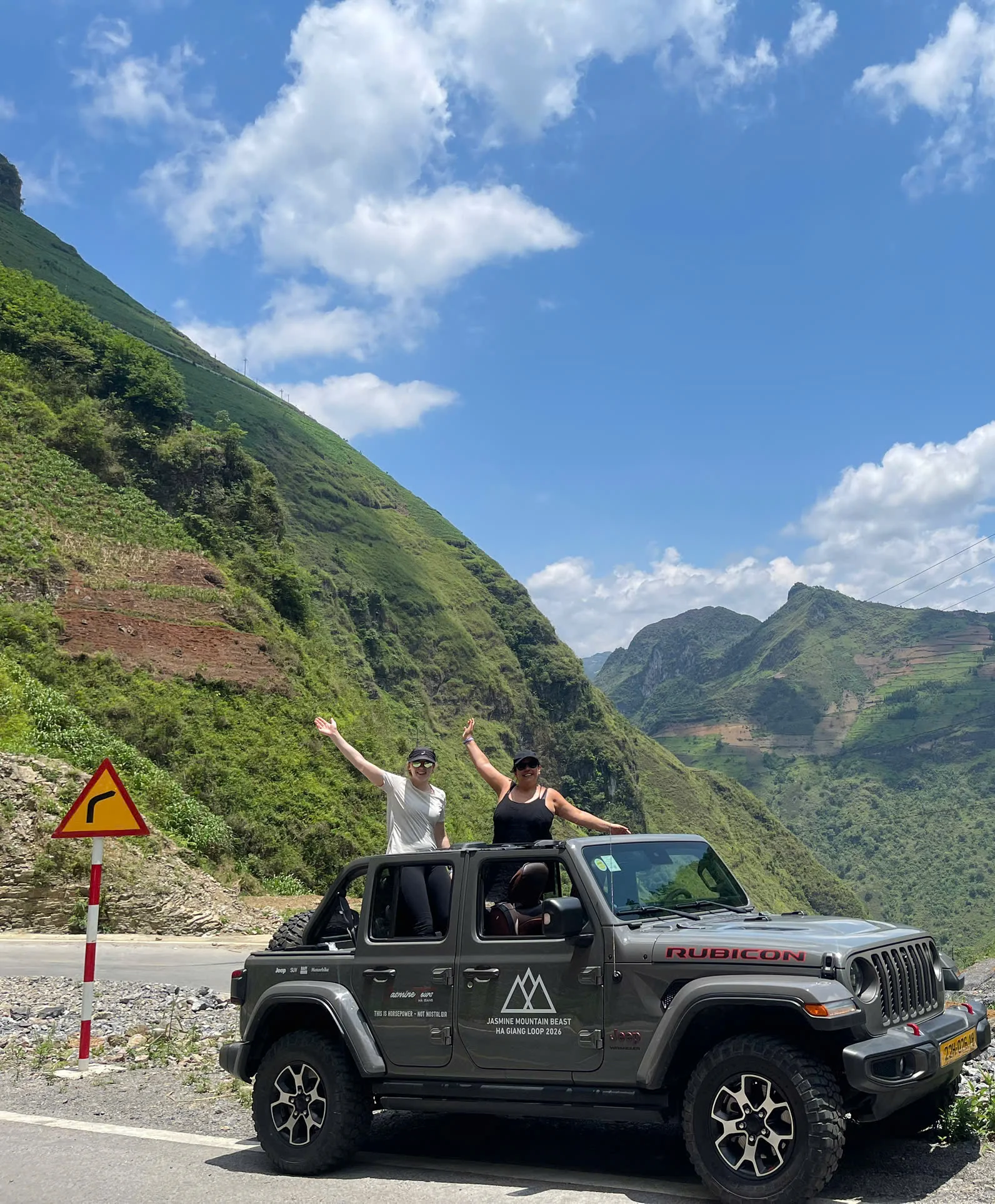A narrow paved road curving between steep karst walls on the Ha Giang Loop, with a yellow sharp-bend warning sign at the roadside, June 2026
