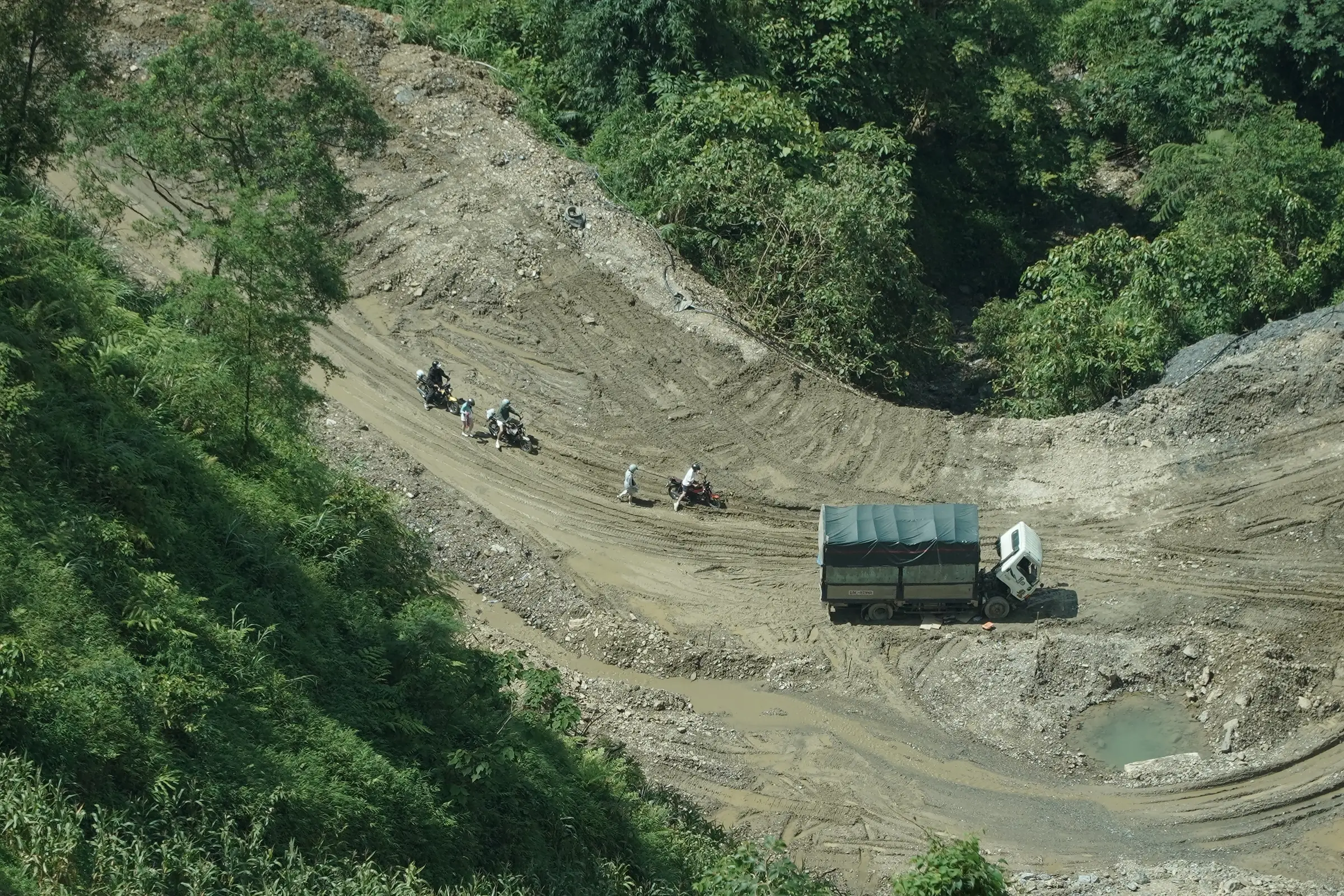 Motorbike riders picking their way across a churned-up landslide repair section beside a parked lorry on the Ha Giang Loop