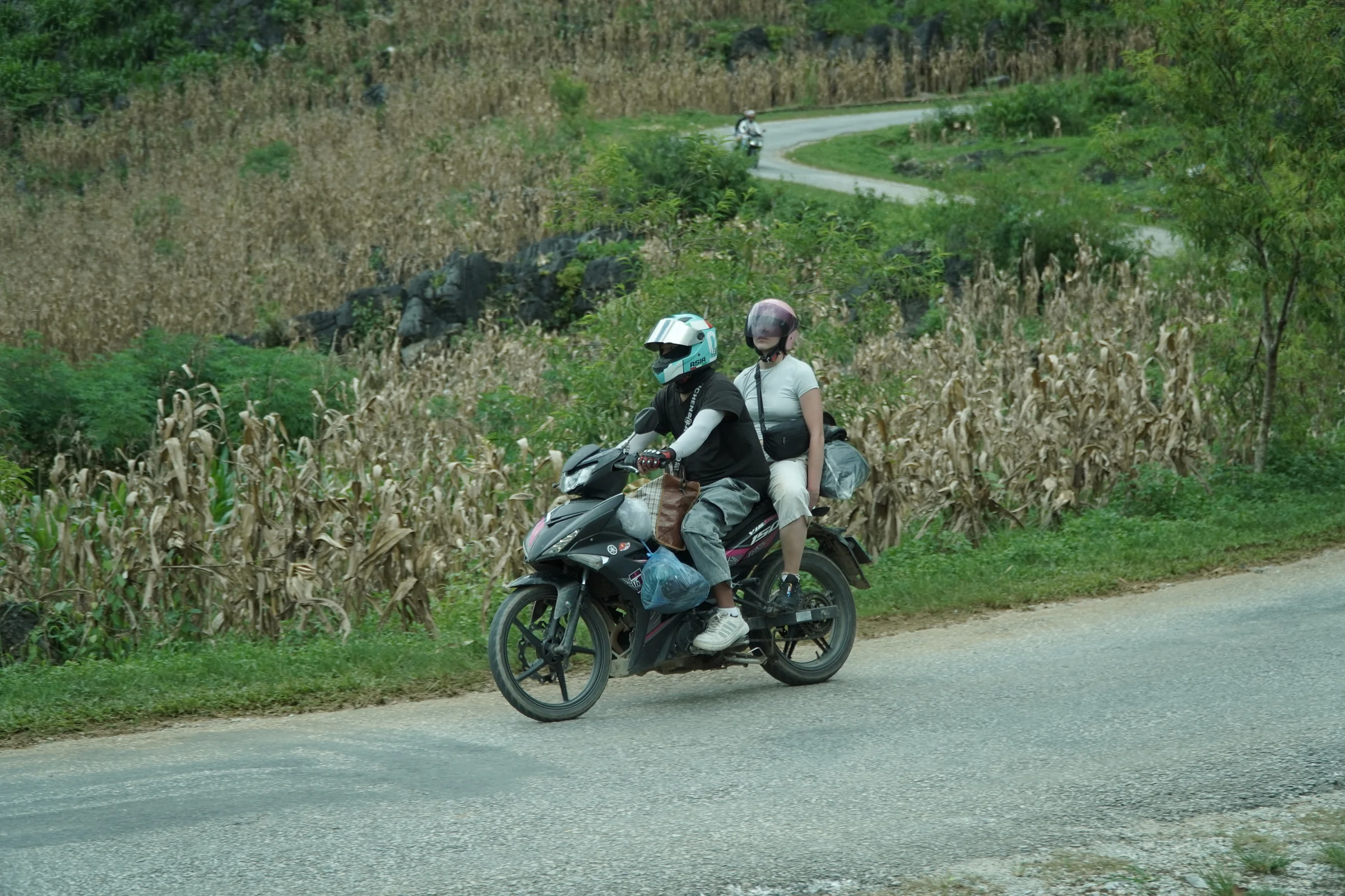 Two people riding two-up on one motorbike on a Ha Giang Loop road, the passenger sitting behind the rider with luggage strapped on